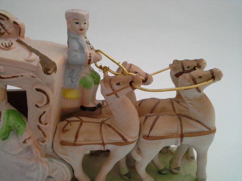 May include: A porcelain figurine of a horse-drawn carriage with a driver in a blue and green outfit. The carriage is pink and white with gold accents. The horses are brown and white.