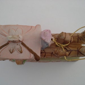 May include: A pink and brown ceramic figurine of a carriage pulled by two horses. The carriage has a floral design and the horses are detailed with brown and white markings. The figurine is tied together with a gold wire.