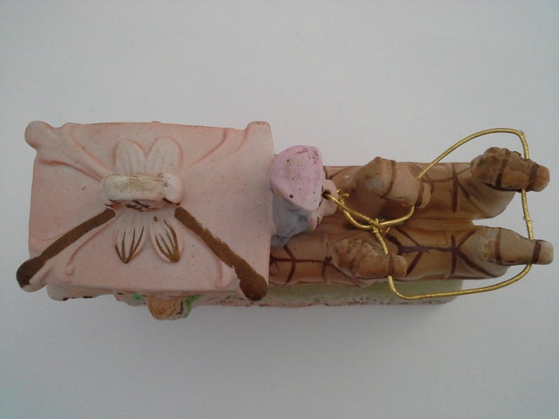 May include: A pink and brown ceramic figurine of a carriage pulled by two horses. The carriage has a floral design and the horses are detailed with brown and white markings. The figurine is tied together with a gold wire.