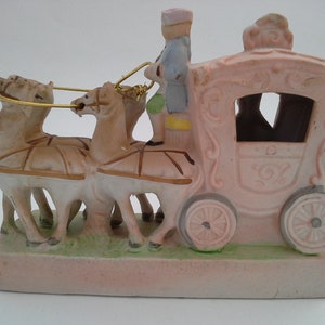 May include: A porcelain figurine of a pink carriage pulled by three horses with a driver in a blue and pink uniform. The carriage has gold accents and a detailed design.