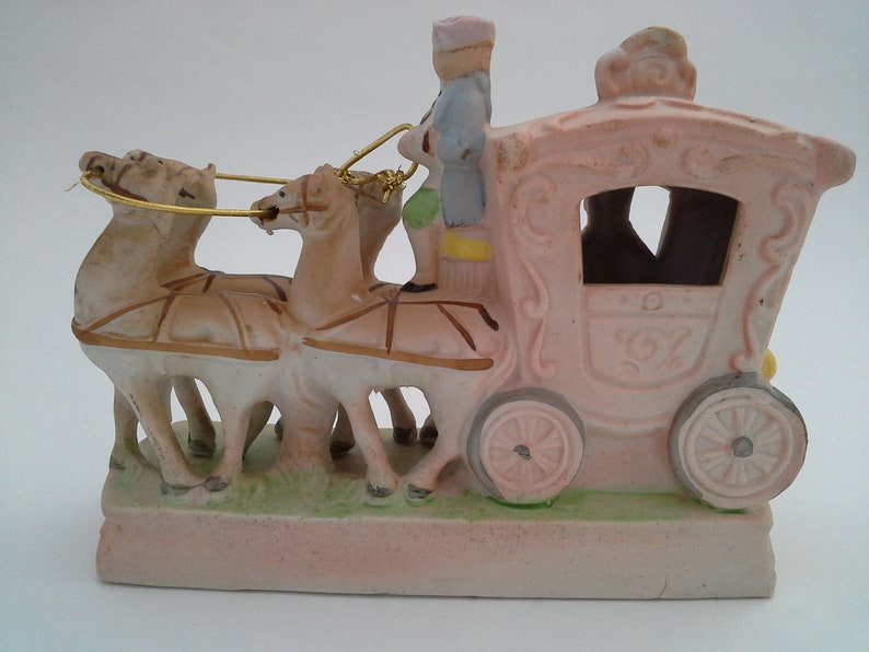 May include: A porcelain figurine of a pink carriage pulled by three horses with a driver in a blue and pink uniform. The carriage has gold accents and a detailed design.