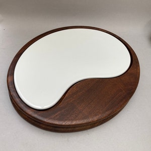 Può includere: Un vassoio da portata bicolore con un inserto in ceramica bianco a forma di rene inserito in una base di legno marrone scuro. La base in legno ha una forma arrotondata e organica. Il vassoio è progettato per servire cibo.
