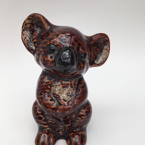May include: A brown ceramic koala ornament with a glossy finish. The koala is in a seated pose with its arms clasped. The ears have a textured, lighter brown pattern. The nose is black.