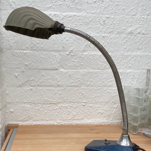 May include: An antique desk lamp with a flexible, silver-coloured neck and a blue base. The lamp shade is a ridged, grey colour. The lamp is on a wooden surface with a white brick wall in the background.