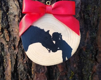 May include: A wooden ornament with a silhouette of a girl kissing a horse. The ornament is decorated with a red bow.
