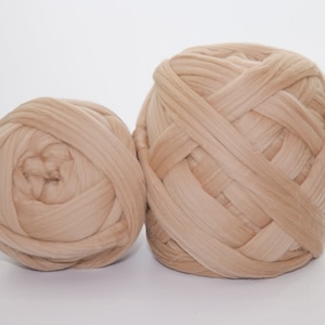 May include: Two beige yarn balls are displayed against a white background. The yarn is thick and appears to be a chunky knit. The balls are different sizes, with one larger than the other. The yarn has a soft, textured appearance.