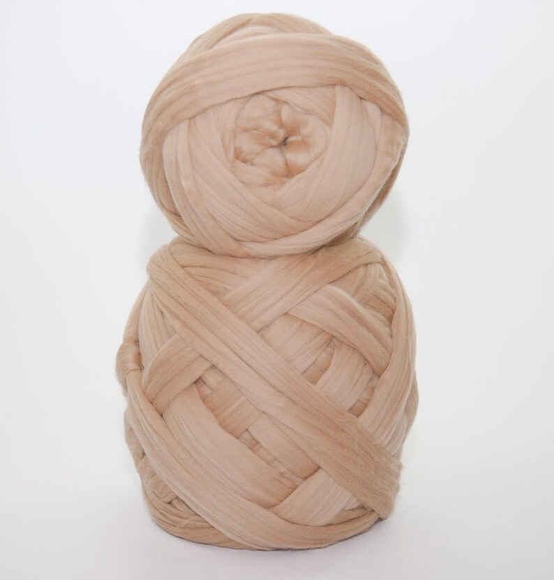 May include: Two large balls of beige yarn are stacked on top of each other against a white background. The yarn is thick and appears to be made of a soft material. The yarn is tightly wound, creating a textured surface.