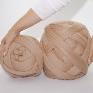 May include: Two large, beige yarn balls. The yarn is thick and appears to be a chunky knit. The balls are held by hands, suggesting their size and texture. Ideal for crafting projects.