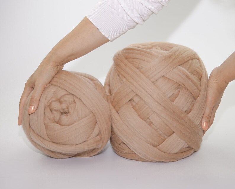 May include: Two large, beige yarn balls. The yarn is thick and appears to be a chunky knit. The balls are held by hands, suggesting their size and texture. Ideal for crafting projects.