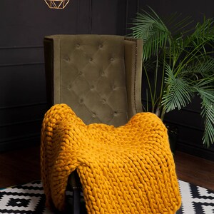 May include: A mustard yellow chunky knit blanket draped over a button-tufted olive green armchair. The blanket's thick weave is visible. Decorative geometric pendant lights hang above. A houseplant is in the background.