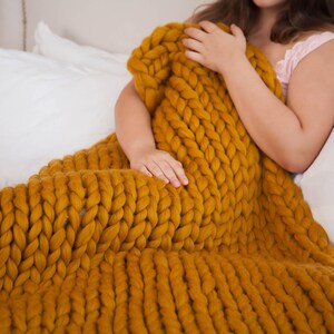 May include: Mustard yellow chunky knit blanket. The blanket is made of thick yarn and has a large, textured weave. The blanket is draped over a bed with white pillows. The blanket is approximately 60 inches wide and 70 inches long.