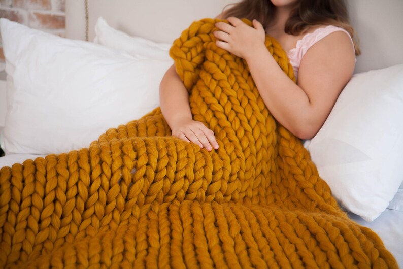 May include: Mustard yellow chunky knit blanket. The blanket is made of thick yarn and has a large, textured weave. The blanket is draped over a bed with white pillows. The blanket is approximately 60 inches wide and 70 inches long.