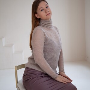 May include: A light brown turtleneck sweater and a mauve satin skirt. The sweater has long sleeves and a semi-sheer appearance. The skirt is knee-length and has a smooth, shiny texture. The person is seated on a gold chair.