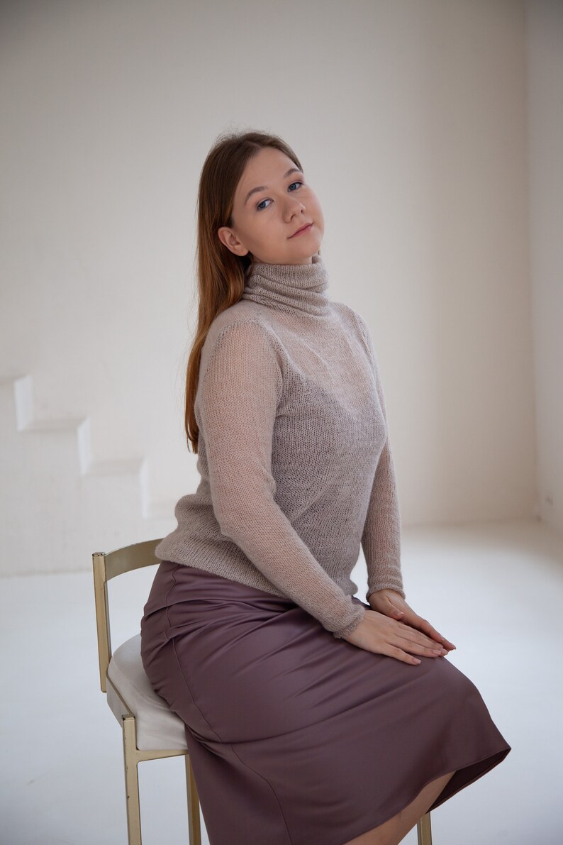 May include: A light brown turtleneck sweater and a mauve satin skirt. The sweater has long sleeves and a semi-sheer appearance. The skirt is knee-length and has a smooth, shiny texture. The person is seated on a gold chair.