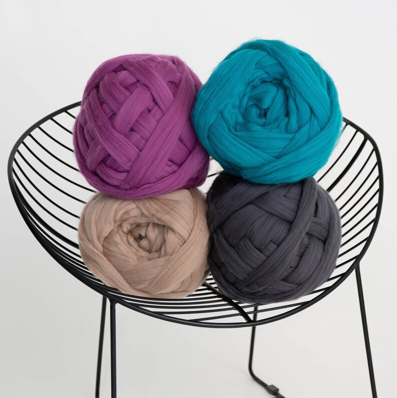 May include: Four large balls of chunky yarn in shades of purple, teal, beige, and charcoal grey are stacked on a black metal wire chair. The yarn appears soft and thick, ideal for creating blankets or other textile projects.