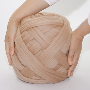 May include: A large ball of beige-colored chunky knit yarn. The yarn is held by two hands, showcasing its size and texture. The yarn appears soft and is suitable for various crafting projects.
