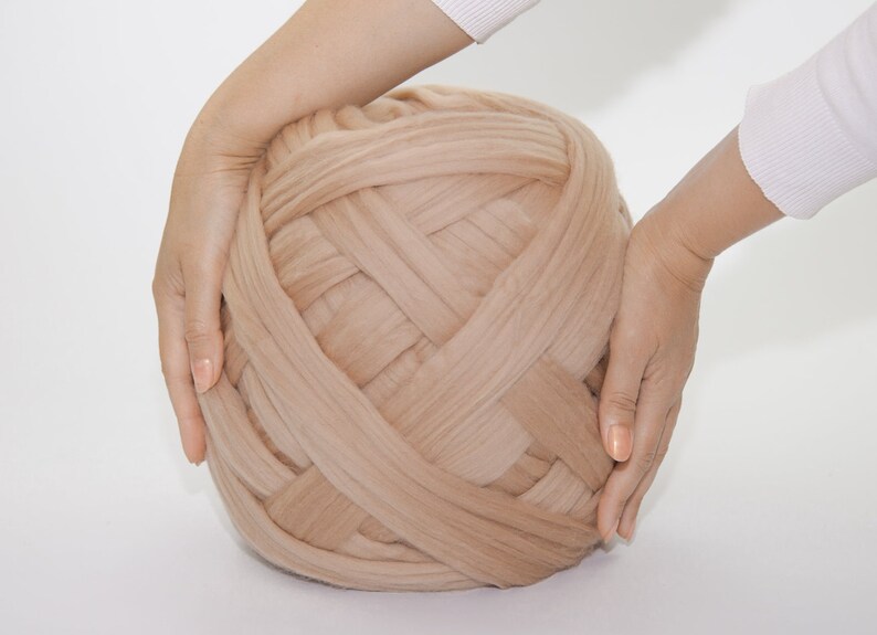 May include: A large ball of beige-colored chunky knit yarn. The yarn is held by two hands, showcasing its size and texture. The yarn appears soft and is suitable for various crafting projects.