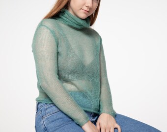 May include: A sheer, seafoam green turtleneck sweater with long sleeves. The sweater is paired with light blue denim jeans. The model is seated, with a neutral background.