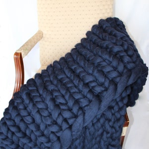 May include: A navy blue chunky knit blanket draped over a beige upholstered chair. The blanket is made of thick, braided yarn, creating a textured, cozy appearance. The blanket's deep blue color contrasts with the chair's neutral tone, suggesting a home decor item.