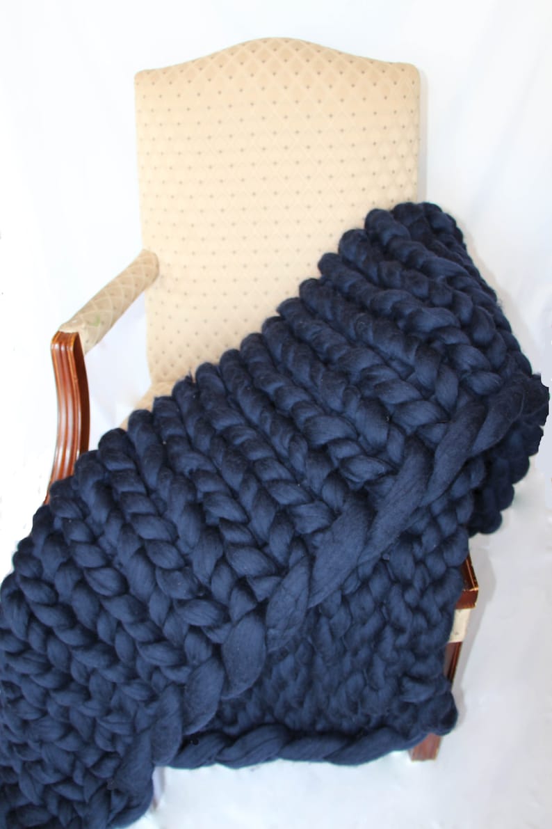 May include: A navy blue chunky knit blanket draped over a beige upholstered chair. The blanket is made of thick, braided yarn, creating a textured, cozy appearance. The blanket's deep blue color contrasts with the chair's neutral tone, suggesting a home decor item.