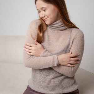 May include: A light beige, sheer turtleneck sweater with long sleeves. The sweater is paired with a mauve satin skirt. The model is sitting on a light-colored couch.