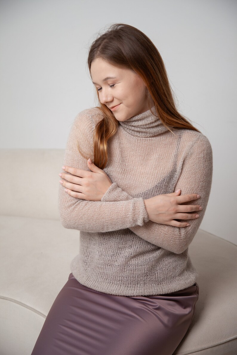 May include: A light beige, sheer turtleneck sweater with long sleeves. The sweater is paired with a mauve satin skirt. The model is sitting on a light-colored couch.