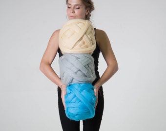 May include: Three large balls of yarn in cream, gray, and turquoise blue are stacked on top of each other. The yarn appears to be thick and chunky, suitable for arm knitting or other large-scale projects. The balls are held by a person.