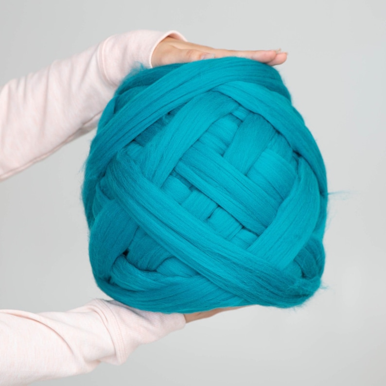 May include: A large ball of teal-coloured super chunky merino wool yarn. The yarn is held by a person with light pink sleeves. The thick yarn is woven in a basket-weave pattern, creating a textured appearance. This yarn is suitable for arm knitting and other craft projects.