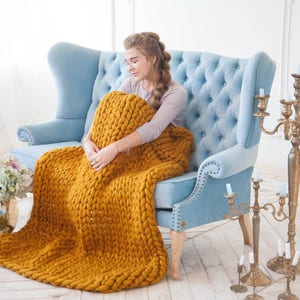 May include: A mustard yellow chunky knit blanket draped over a light blue tufted armchair. The blanket is thick and textured, with visible large stitches. The room has a bright, airy feel, with a gold candelabra and floral arrangements.