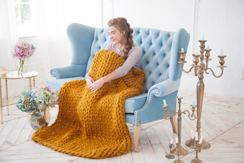 May include: A mustard yellow chunky knit blanket draped over a light blue tufted armchair. The blanket is thick and textured, with visible large stitches. The room has a bright, airy feel, with a gold candelabra and floral arrangements.