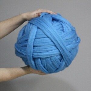 May include: A large ball of light blue chunky knit yarn. The yarn is held by two hands, with one hand on top of the ball. The yarn appears to be made of thick, soft fibers, suitable for arm knitting or other large-scale projects.