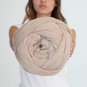 May include: A large ball of beige yarn, held in front of a person. The yarn appears to be thick and soft, suitable for chunky knitting or arm knitting projects. The neutral colour would complement various home decor styles.