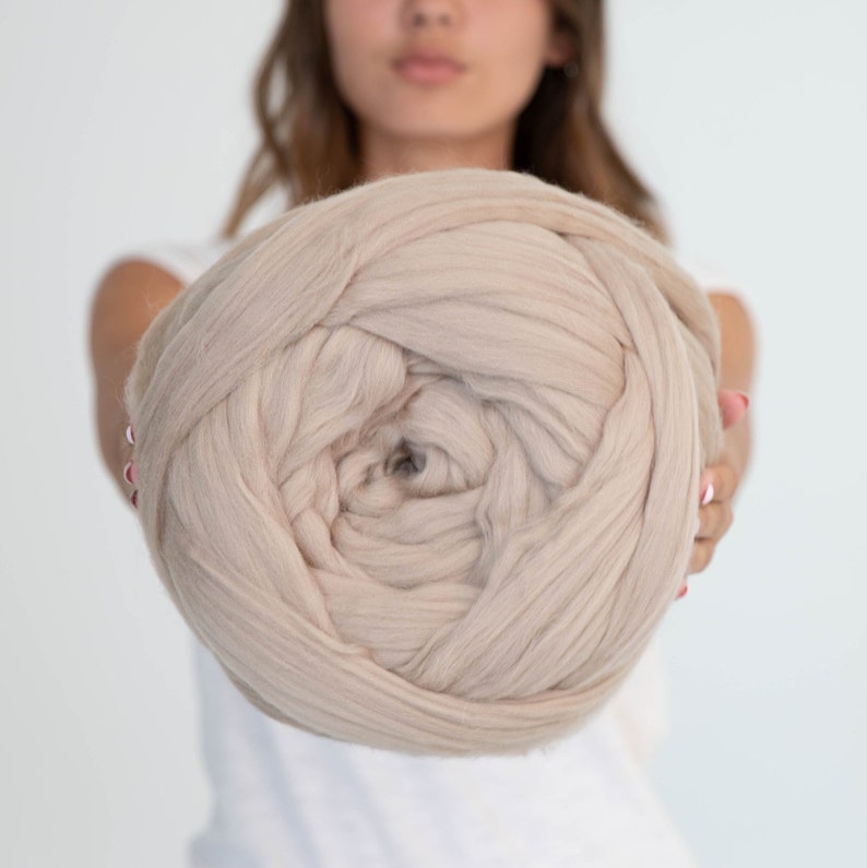 May include: A large ball of beige yarn, held in front of a person. The yarn appears to be thick and soft, suitable for chunky knitting or arm knitting projects. The neutral colour would complement various home decor styles.