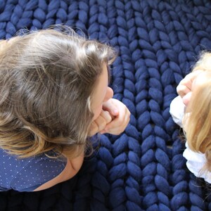 May include: A close-up shot of a large, chunky knit blanket in a deep navy blue. Two people are seen lying on the blanket, their faces close together. The blanket's texture is prominent, suggesting a cozy and warm item.