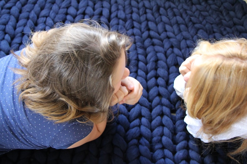 May include: A close-up shot of a large, chunky knit blanket in a deep navy blue. Two people are seen lying on the blanket, their faces close together. The blanket's texture is prominent, suggesting a cozy and warm item.