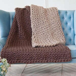 May include: Two chunky knit blankets in brown and beige are draped over a light blue upholstered armchair. The blankets have a thick, textured appearance, with visible stitches. The brown blanket is folded at the bottom, while the beige one is spread out.