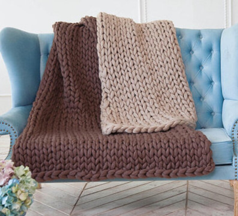 May include: Two chunky knit blankets in brown and beige are draped over a light blue upholstered armchair. The blankets have a thick, textured appearance, with visible stitches. The brown blanket is folded at the bottom, while the beige one is spread out.