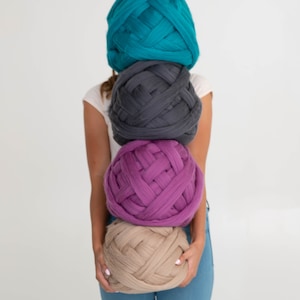 May include: Four large balls of chunky yarn in teal, grey, purple, and beige are stacked in front of a person. The yarn is thick and woven, suggesting a craft project. The person is wearing a white top and blue jeans.