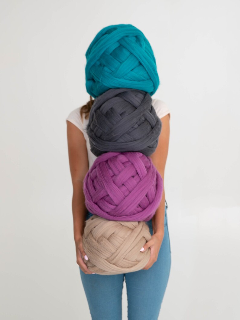 May include: Four large balls of chunky yarn in teal, grey, purple, and beige are stacked in front of a person. The yarn is thick and woven, suggesting a craft project. The person is wearing a white top and blue jeans.