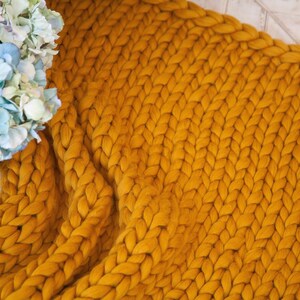 May include: Mustard yellow chunky knit blanket. The blanket is made of thick yarn and has a braided pattern. A vase of blue and purple hydrangeas is in the background. The blanket is draped over a surface.