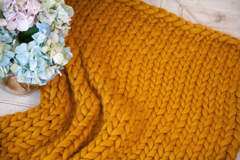 May include: Mustard yellow chunky knit blanket. The blanket is made of thick yarn and has a braided pattern. A vase of blue and purple hydrangeas is in the background. The blanket is draped over a surface.