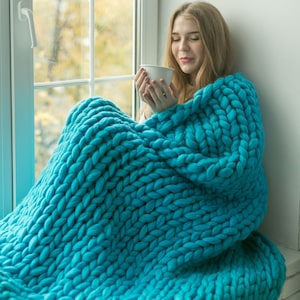 May include: A person wrapped in a large, chunky-knit teal blanket, holding a white mug. The blanket is made of thick yarn, creating a cosy and textured appearance. The scene is set near a window with an out-of-focus background.