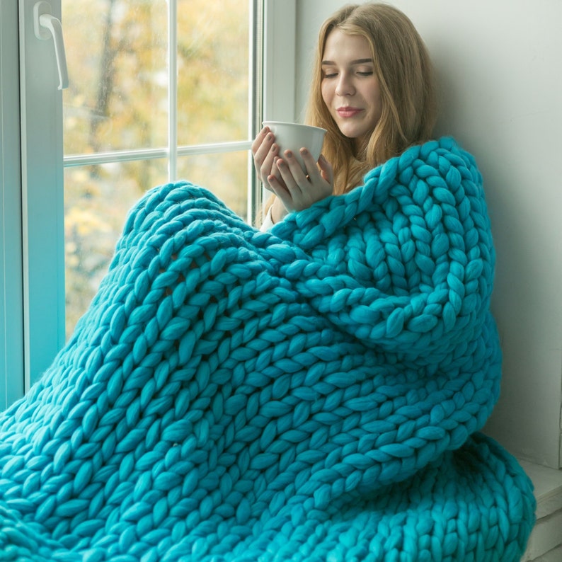 May include: A person wrapped in a large, chunky-knit teal blanket, holding a white mug. The blanket is made of thick yarn, creating a cosy and textured appearance. The scene is set near a window with an out-of-focus background.