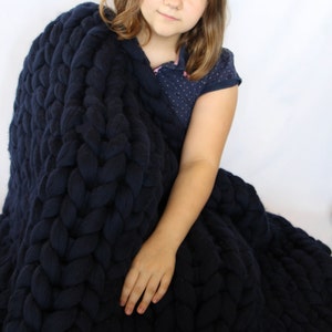 May include: A navy blue chunky knit blanket. The blanket is made of thick, soft yarn and has a large, braided texture. The blanket is draped over a person, creating a cozy and inviting look. The blanket is a solid color and appears to be very warm.