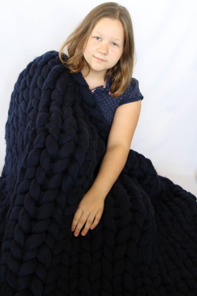 May include: A navy blue chunky knit blanket. The blanket is made of thick, soft yarn and has a large, braided texture. The blanket is draped over a person, creating a cozy and inviting look. The blanket is a solid color and appears to be very warm.