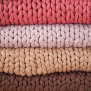 May include: A stack of chunky knit blankets in various colors. The blankets are in shades of beige, coral, light pink, tan, brown, and mustard yellow. The thick yarn is woven in a large, visible stitch pattern, creating a textured surface.