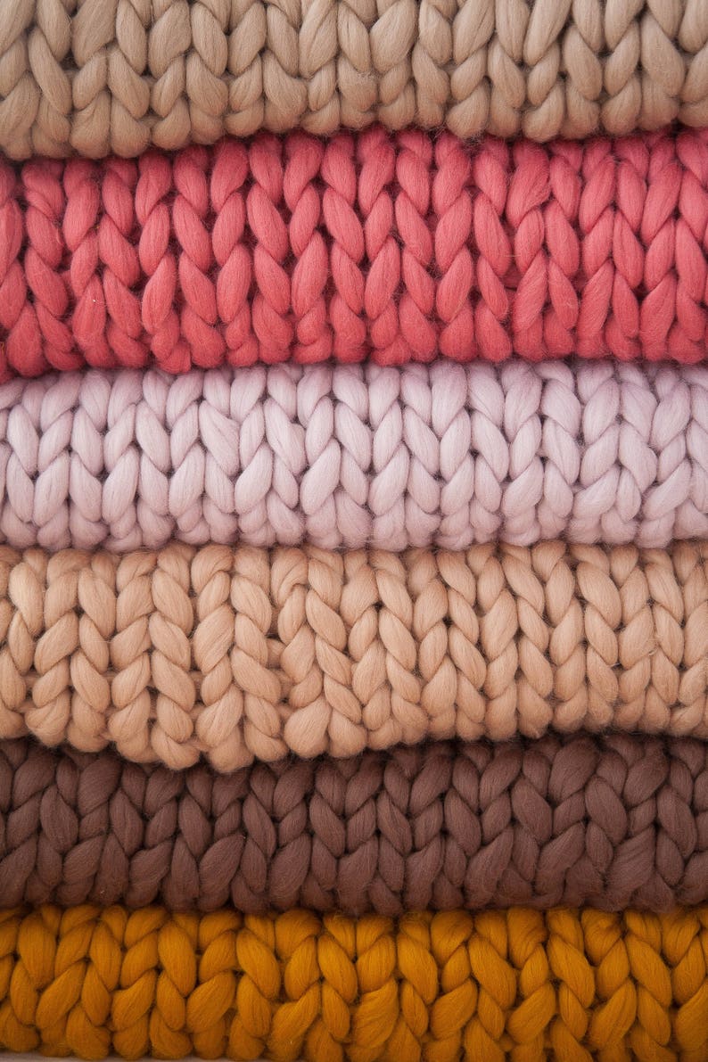 May include: A stack of chunky knit blankets in various colors. The blankets are in shades of beige, coral, light pink, tan, brown, and mustard yellow. The thick yarn is woven in a large, visible stitch pattern, creating a textured surface.