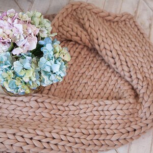 May include: A chunky knit blanket in a neutral beige color is draped on a light-colored wooden surface. A glass vase holds a bouquet of hydrangeas in shades of white, pink, blue, and green, adding a touch of color.