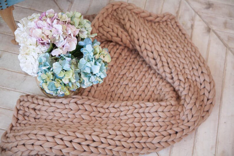 May include: A chunky knit blanket in a neutral beige color is draped on a light-colored wooden surface. A glass vase holds a bouquet of hydrangeas in shades of white, pink, blue, and green, adding a touch of color.