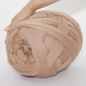 May include: A large ball of beige yarn, tightly woven and textured, is held by a hand. The yarn appears soft and thick, suitable for chunky knit projects. The neutral color makes it versatile for various crafting needs.